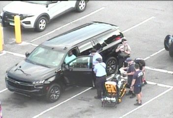 Several individuals, some appearing to be emergency personnel, attending to a stretcher next to a black SUV with its door open.