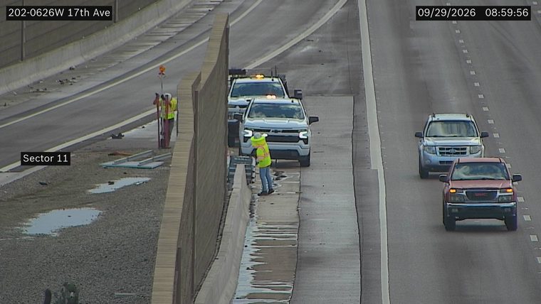 Two workers in high-visibility vests and hard hats are present near construction materials and a tow truck with flashing amber lights.