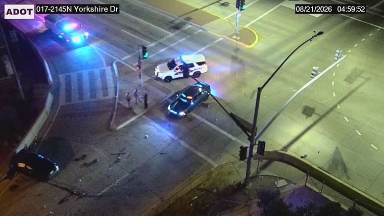 Two police vehicles with lights on and three individuals standing near a dark sedan in the intersection