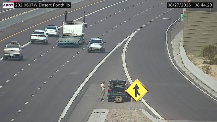 Person standing next to a car with an open trunk and a spare tire on the ground, on the right shoulder