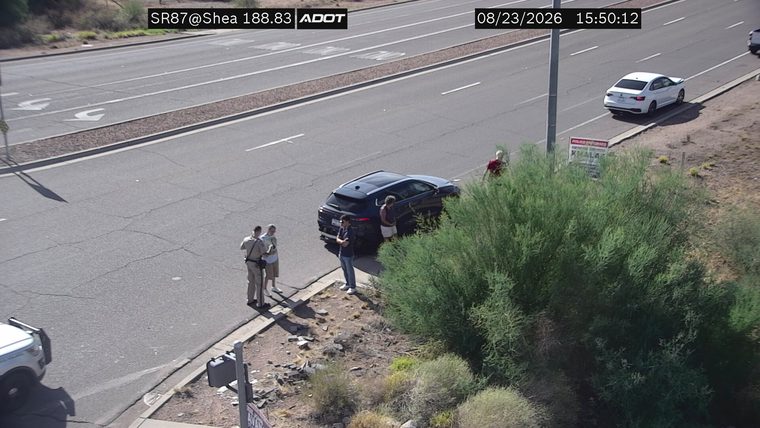 Two individuals in uniform, appearing to be law enforcement, are standing near a dark SUV on the shoulder of the road. A white sedan is visible further down the road.