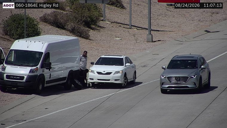 White sedan with a person attending to the front passenger side tire, next to a white van on the right shoulder