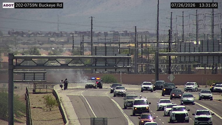 Police car with red and blue lights on the right shoulder, with two individuals standing nearby and smoke or steam rising from the area.