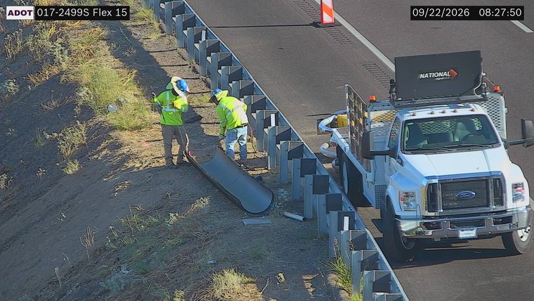 Two workers with high-visibility vests and hard hats working on a drainage pipe next to the highway with a service truck present