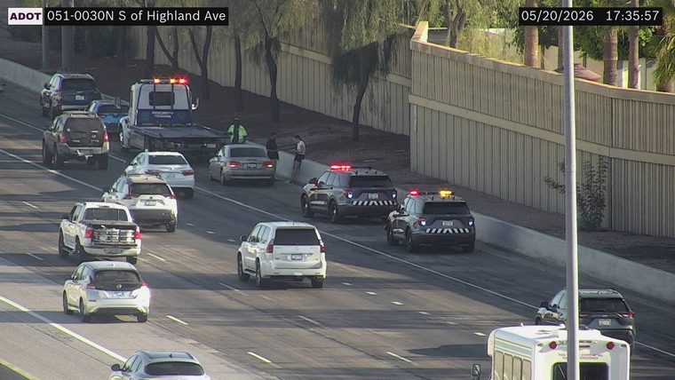 Two police vehicles with flashing lights on the right shoulder and a tow truck with a worker on the left shoulder
