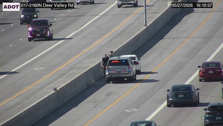 Police vehicle with emergency lights on and two people standing on the left shoulder next to a stopped white sedan