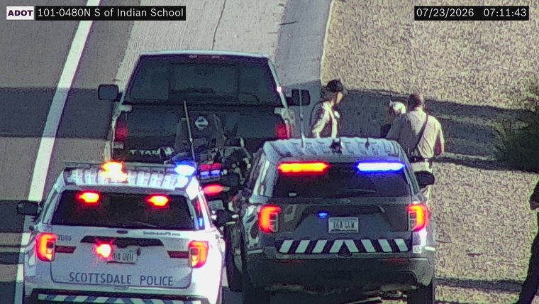 Two police vehicles with lights on and three individuals near a pickup truck on the right shoulder