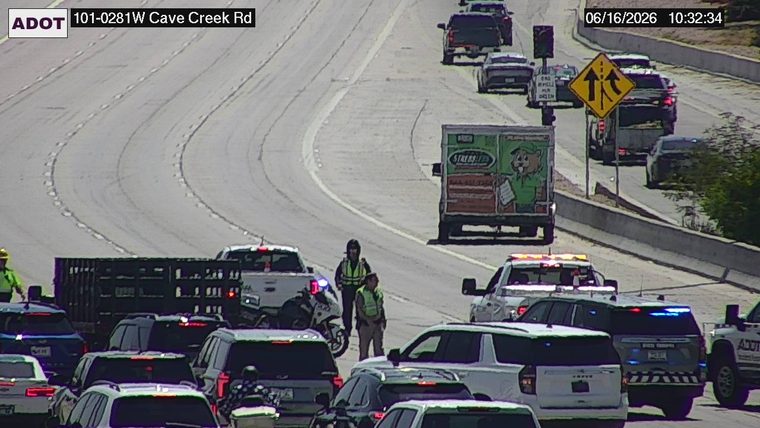 Two individuals in high-visibility vests and a police motorcycle are present in the roadway among stopped vehicles.