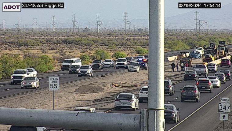 Multiple vehicles and construction equipment in the roadway, with several individuals standing nearby