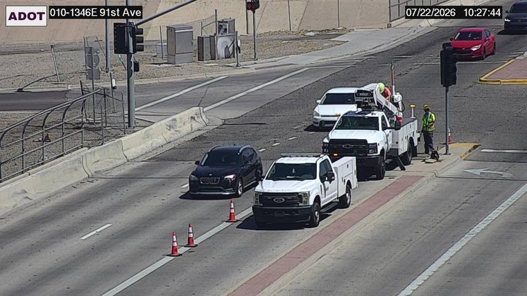 Two workers in high-visibility vests and a utility truck with a crane are on the right shoulder, with traffic cones in the lane.