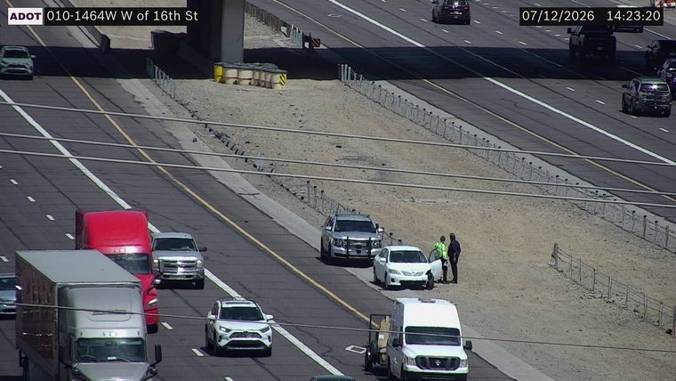 Two individuals standing next to a white sedan on the right shoulder, with a silver SUV stopped behind it.