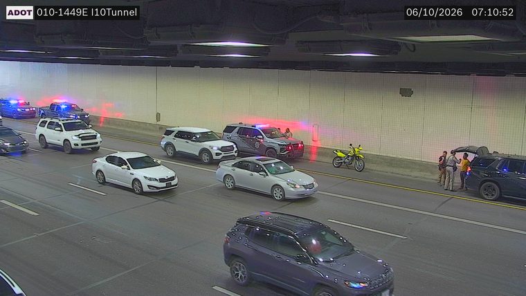 Two police officers and a civilian interacting near a dark SUV with its trunk open and a motorcycle parked nearby on the right shoulder