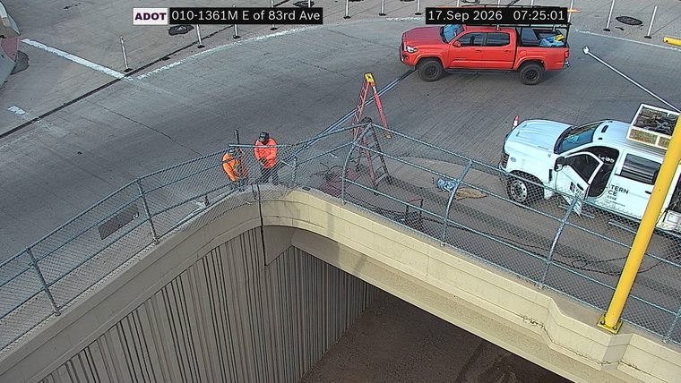 Two workers in orange vests and hard hats near a ladder and equipment on the roadway
