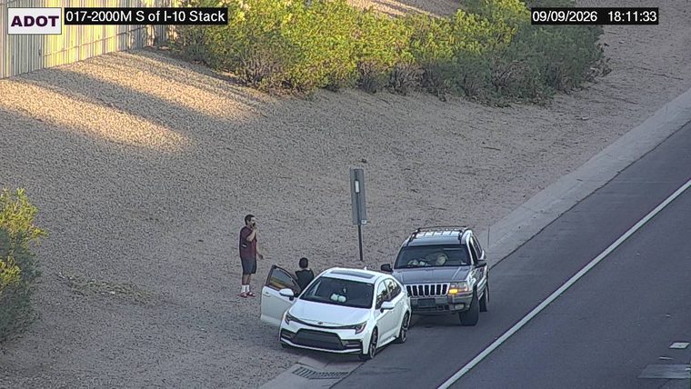 Two persons standing near a white car with its door open, and a dark SUV parked nearby, all off the travel lanes
