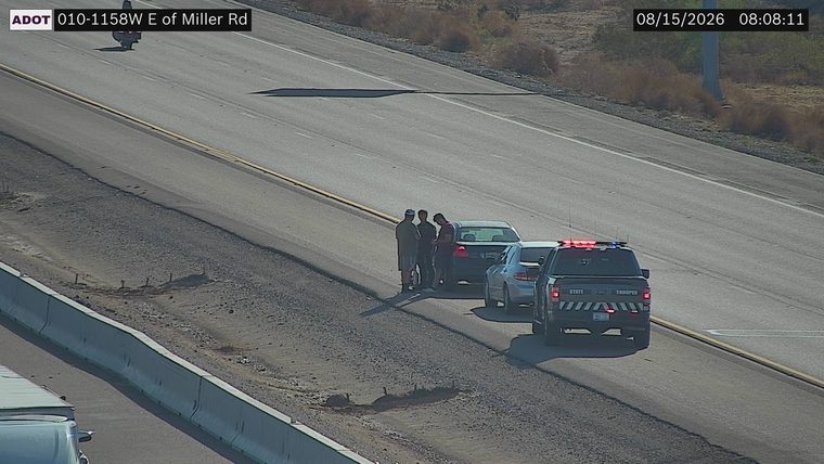 State Trooper vehicle with red and blue lights on the right shoulder next to a stopped car with three individuals standing nearby
