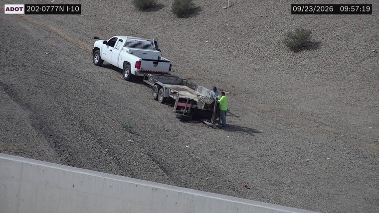 Two workers near a trailer with a white pickup truck attached, off the roadway