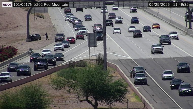 Crashed vehicle on the left dirt shoulder against a pole base with a person standing nearby, and two vehicles stopped on the right shoulder with a person standing outside