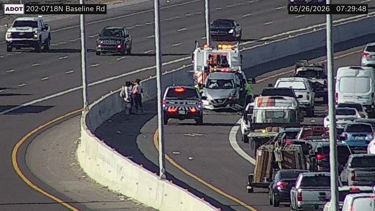 Crash scene on the left shoulder with a damaged silver SUV, emergency response vehicles with flashing lights, and people standing by the concrete barrier