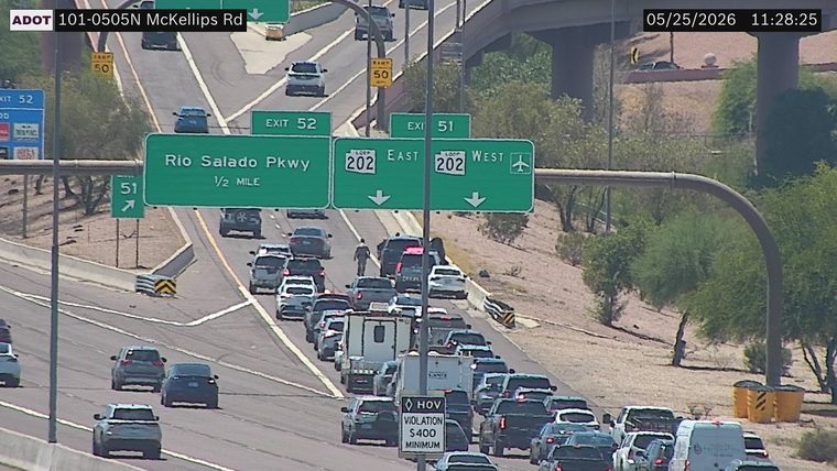 Pedestrian walking on the roadway between lanes of traffic under the Loop 202 East sign