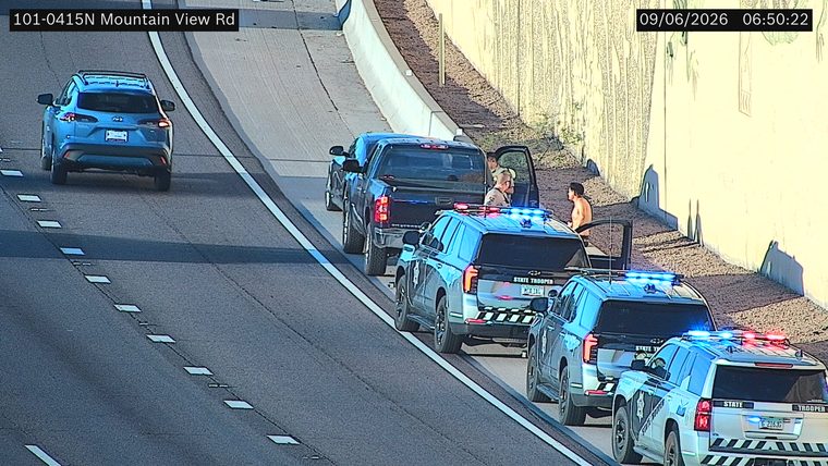Three state trooper vehicles with lights on, stopped on the right shoulder next to a pickup truck with its door open and two individuals present