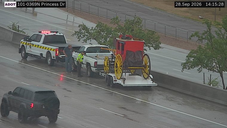 Tow truck with red flashing lights assisting a white pickup truck towing a stagecoach on a trailer, all on the right shoulder