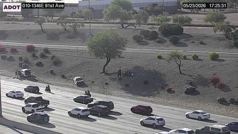 White SUV on the left shoulder with its rear hatch open and a person standing nearby, with another pickup truck and a group of people on the roadside embankment