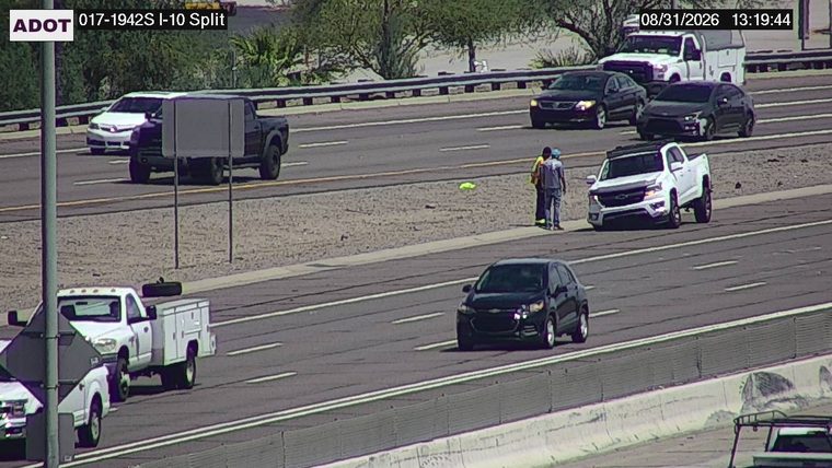 Two individuals in high-visibility vests standing next to a white pickup truck on the right shoulder
