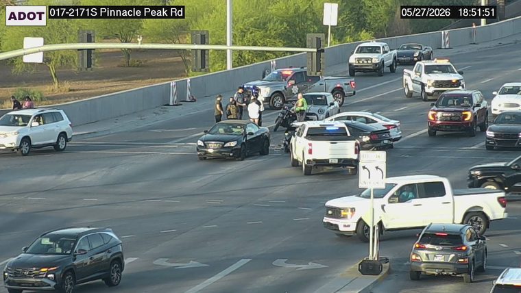 Multiple police vehicles with emergency lights active and several people standing on the roadway near a stopped black convertible