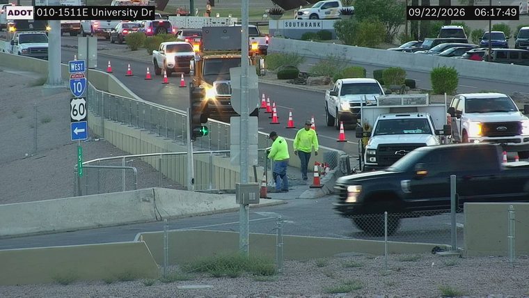 Two workers in high-visibility vests near traffic cones and equipment on the right side of the roadway