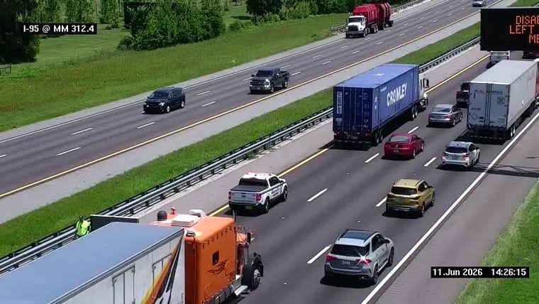 Person in high-visibility vest near disabled vehicle on highway shoulder.