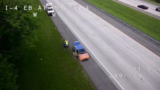 Two people and a car are stopped on the shoulder, one person in a safety vest.