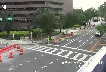 Two workers in high-visibility vests are present near traffic cones on the roadway.