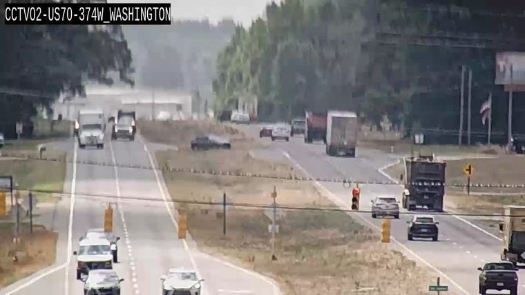 Person in orange vest directing traffic near a dump truck and cars.