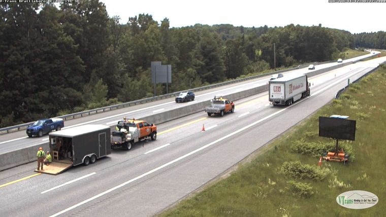 Two workers and a trailer are on the shoulder of the highway with traffic cones present.