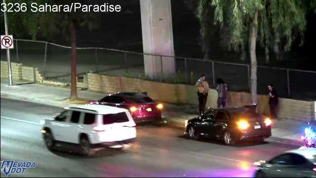 Police officer standing on the sidewalk next to a stopped black sedan, with emergency lights visible on the right