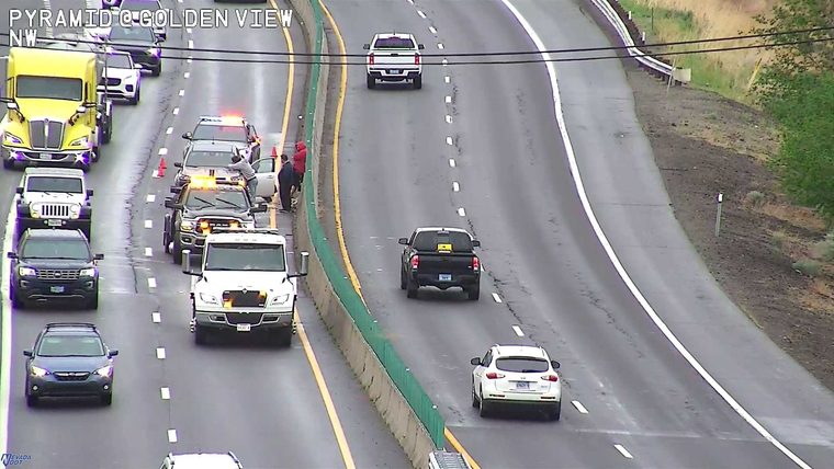 Police car with flashing lights, tow trucks, and people standing next to a vehicle on the left shoulder/median