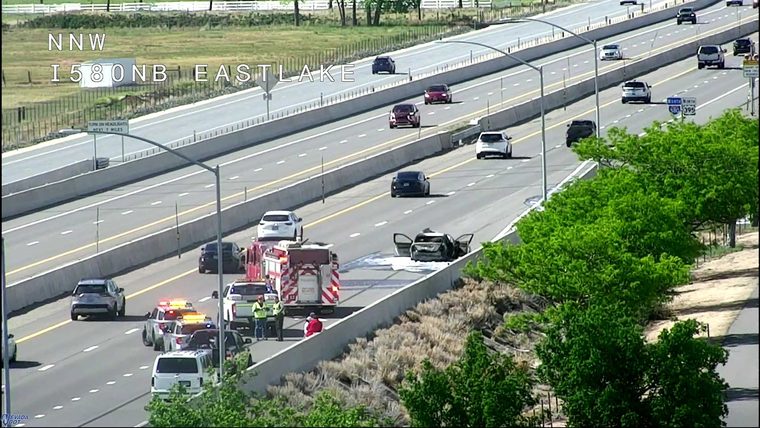 Fire truck and multiple police cars with lights on near a burning car in the right lane
