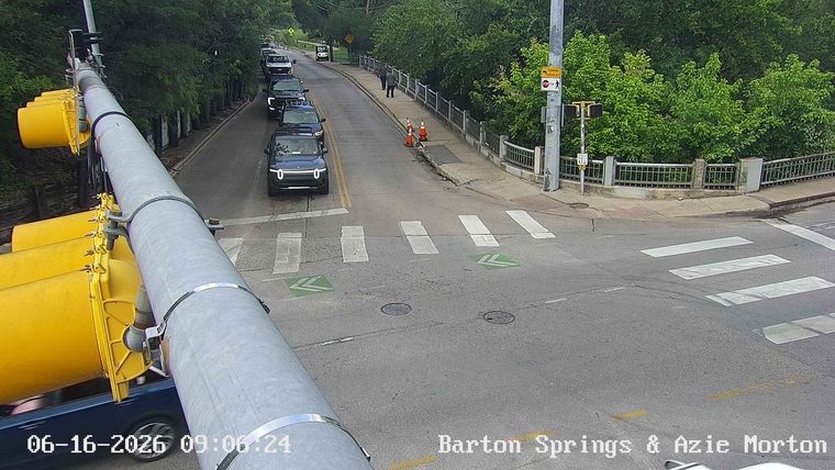Traffic cones and two people on the side of the road near a bridge.