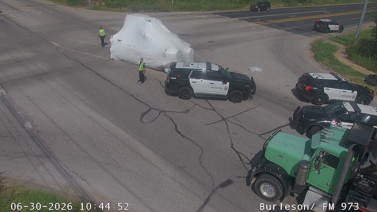 Police cars and two individuals in high-visibility vests near a large object covered in white tarp in the intersection