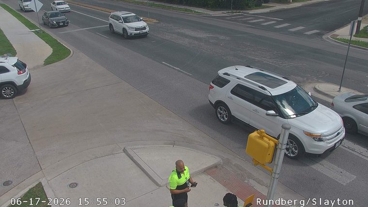 Two individuals in high-visibility vests are standing near a traffic light pole.