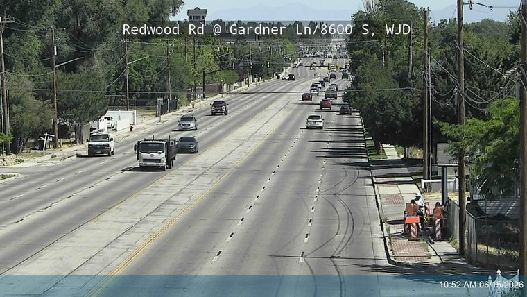 Two workers in orange vests with cones are present on the roadside.