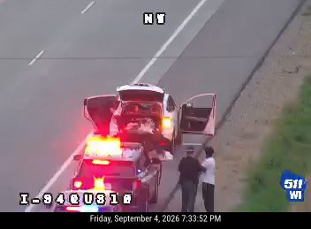 Police car with red and blue lights on the right shoulder with two people standing nearby