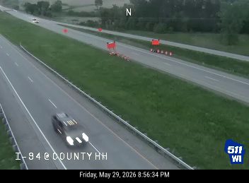 Police vehicle on shoulder with emergency lights active, traffic cones present.