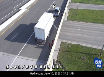 Two people appear to be responders next to a stopped truck on the highway shoulder.