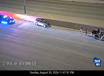 Police car with red and blue lights on the left shoulder with two people standing near it